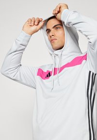 Hellgrauer Hoodie mit pinkem diagonalen Streifen, drei schwarzen Streifen an den Ärmeln, geripptem Textur, weißen Kordeln und einer taillierten Passform.