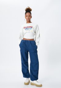 Witte cropped sweatshirt met kleurrijk grafisch ontwerp; blauwe losse broek; witte chunky schoenen met een gestructureerde zool. Ronde hoepel oorbellen.