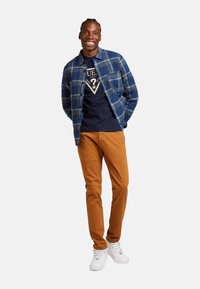 Camisa de cuadros azul sobre una camiseta navy, combinada con pantalones naranjas y zapatillas blancas. El modelo está de pie con las manos en los bolsillos, sonriendo.
