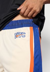 Krēma krāsas basketbola šorti ar zilām un oranžām sānu joslām, ar izšūtu "NYC" logo un elastīgu jostasvietu.