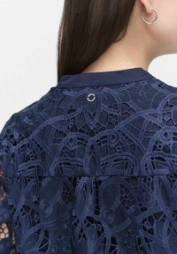 Granatowa koronkowa bluzka z misternymi wzorami kwiatowymi, przezroczystą teksturą oraz małym okrągłym metalowym akcentem na tylnym dekolcie.