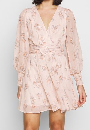 Kobieta w jasnoróżowej, kwiecistej sukience kopertowej z długimi bufkami na rękawach i szerokim paskiem z okrągłą klamrą w talii.