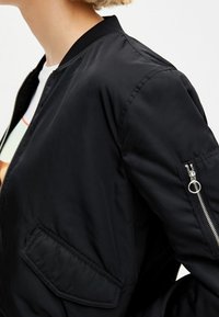 Personne portant une veste bomber noire montrant le haut du torse et le bras avec une poche avant et une poche zippée sur la manche.