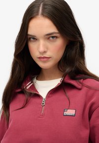 Bordeaux rode sweatvest met ritssluiting en een witte kraag, voorzien van een marineblauw 'Superdry'-label met een Amerikaanse vlag. De stof lijkt glad en comfortabel.