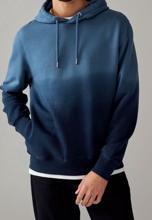 Homme portant un sweat à capuche bleu dégradé avec cordons, t-shirt blanc en dessous et pantalon noir, debout avec une main dans la poche.