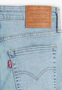Pantalones vaqueros de mezclilla azul claro con una parche trasero de cuero con logo en relieve, detalles de costura sutiles y una etiqueta roja en el bolsillo lateral.
