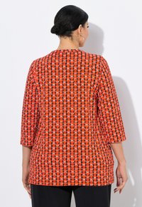 Nainen, jolla on tumma hiusnyppy, pukeutuneena punaisiin ja mustiin kuvioituihin pitkähihaisiin paitoihin ja mustiin housuihin, seisoo valkoista taustaa vasten selkä kameraan.