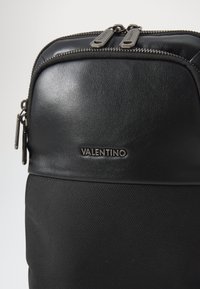 Zaino nero con sezione superiore in pelle e parte inferiore in nylon texturizzato, dotato di doppie cerniere e logo Valentino sulla parte anteriore.