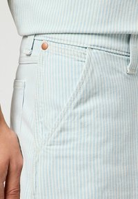 Pantalones de algodón a rayas azul claro con un bolsillo lateral, detalle de botón de cobre y costuras en contraste. Vista en primer plano de la textura de la tela.