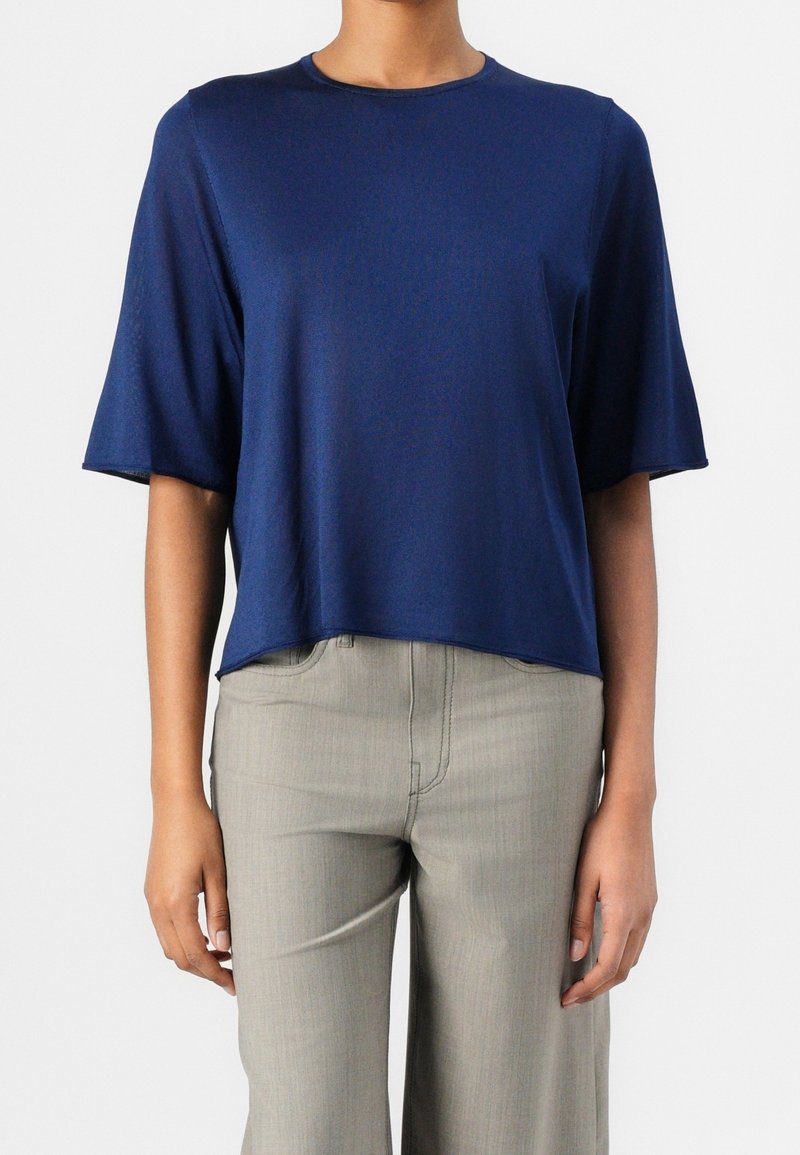 Personne portant un haut bleu marine ample à manches courtes et un pantalon gris clair, debout devant un fond blanc uni.