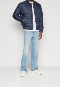 Marinblå puffjacka med quiltat mönster och ribbade muddar, bärs över en grå skjorta; i kombination med ljusblå jeans och vita sneakers.