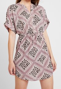 Woman wearing a short-sleeve, pink and black patterned dress with a drawstring waist and V-neckline, standing against a light background.