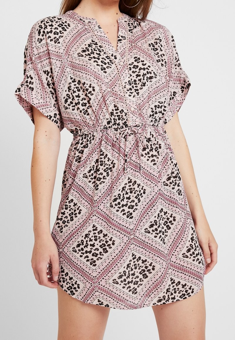 Woman wearing a short-sleeve, pink and black patterned dress with a drawstring waist and V-neckline, standing against a light background.