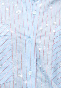 Ανοιχτό μπλε ύφασμα με λεπτές κόκκινες κάθετες ρίγες, διακοσμημένο με διασκορπισμένες διάφανες παγιέτες και λευκά κουμπιά, με γωνιακές λεπτομέρειες στη ραφή.