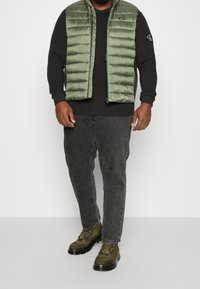 Gilet matelassé olive sur une chemise noire à manches longues, associé à un jean gris foncé et des bottines brunes. Comprend des accents de zipper et de logo.