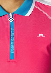 Růžové polokošile s modrým límečkem a zipem. Na přední straně modré logo "J.Lindeberg". Hladká, lehká látka s bílými detaily.