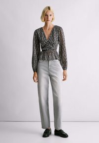 Priehľadná čierna blúzka s potlačou, dlhými rukávmi a pásom s volánmi, v kombinácii so svetlosivými džínsami s rovnými nohavicami a čiernou obuvou.