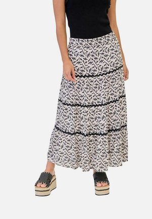 Mujer con un top negro sin mangas, falda larga blanca de varios volantes con patrón de hojas negras y sandalias negras de plataforma con detalles de cuerda.