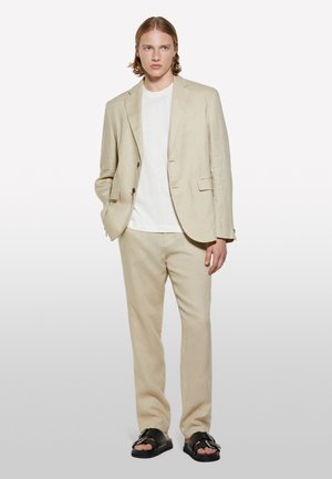 Helles beiges Leinenanzug mit einem maßgeschneiderten Blazer und geraden Hosen, kombiniert mit einem weißen T-Shirt und schwarzen Sandalen. Glatte Textur und klare Linien.