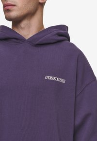 Sudadera oversized morada de algodón, con un bolsillo frontal y un pequeño logo "PEGADOR" en blanco en el lado izquierdo del pecho. Textura suave.