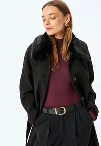 Cappotto di lana nero con colletto di pelliccia, caratterizzato da un design doppiopetto e grandi bottoni, indossato sopra un top bordeaux e pantaloni neri.