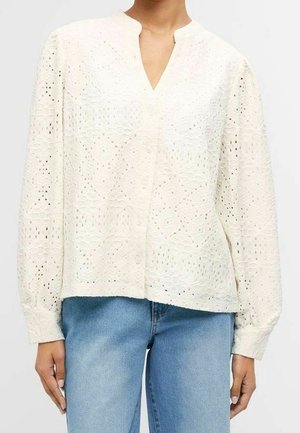 Blouse blanche à manches longues avec motif floral en broderie anglaise, manches légèrement bouffantes et col en V, portée avec un jean denim bleu clair.