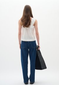 Haut sans manches blanc avec détails texturés, associé à un jean en denim bleu ample. Modèle tenant un sac à bandoulière noir. Fond minimaliste.