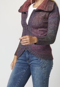 Femme portant un pull en tricot multicolore à fermeture éclair avec un grand col et un jean brodé bleu, debout devant un fond uni.