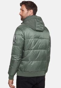 Vadderad grön jacka med huva, med quiltade sektioner, ribbad nederkant och ett runt logopatch på vänster arm. Slät textur.