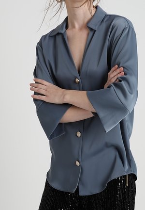 Vrouw draagt een losse blauwe blouse met knopen en driekwart mouwen, armen gekruist, gecombineerd met zwarte paillettenbroek.