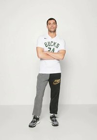 Mies seisoo kädet ristissä, päällä valkoinen Bucks koripallopaita ja mustaharmaat housut, joissa on kultainen Nike-logo, sekä harmaa-mustat lenkkarit.