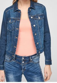 Chaqueta de mezclilla con un tono oscuro, cierres de botón, bolsillos en el pecho y costuras naranjas, combinada con una blusa rosa claro y jeans azules.