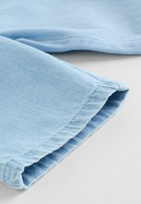 Lyseblåt denimstof foldet ved kanten med synlige syninger på en ensfarvet hvid overflade.