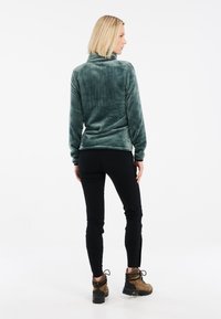 Zelená fleecová mikina s vysokým límcem, dlouhými rukávy a hladkým povrchem. Nosit s černými slim-fit kalhotami a hnědými turistickými botami.