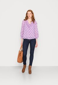 Blusa lavanda com um padrão de motivos roxos escuros, com mangas longas e decote em V, emparelhada com jeans skinny escuros e botas de tornozelo castanhas.