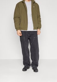 Giubbotto bomber verde oliva con polsini elastici, abbinato a jeans neri larghi con cuciture a contrasto e due tasche frontali. Footwear neri.