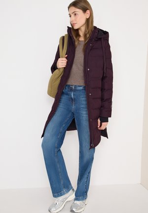 Giovane donna che indossa un piumino viola scuro, jeans blu, sneaker argentate e porta una borsa a tracolla verde oliva, in piedi contro un muro bianco.