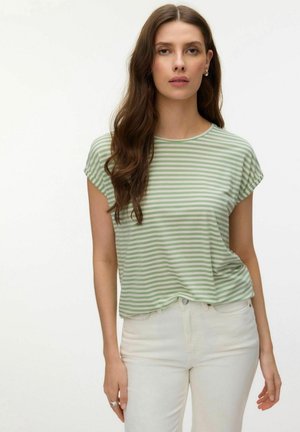 Kvinde med langt brunt hår iført en grøn- og hvidstribet kortærmet skjorte og hvide bukser, stående mod en ensfarvet baggrund.