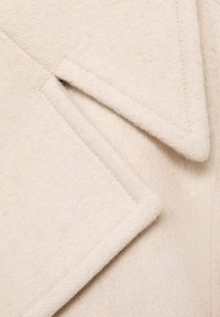Tessuto di un cappotto in lana beige con una texture morbida, caratterizzato da rever inclinati e cuciture a vista, che enfatizzano gli elementi di design a strati.