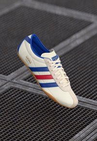 Adidași albi sport cu dungi albastre și roșii, talpă din cauciuc texturat și căptușeală interioară albastră. Prezintă șireturi și branding pe lateral.