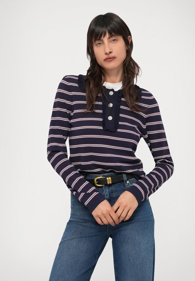 Mornarsko modra oranžno črtasta majica z dolgimi rokavi z nabrano izrezom in gumbe na sprednji strani, združena z modrimi kavbojkami z visokim pasom in črnim pasom.