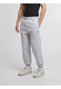 Lichtgrijze sweatpants met een elastische tailleband, zijzakken en zwart geborduurd logo. Gecombineerd met lichtgekleurde sport sneakers.
