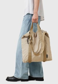 Bolsa de tela beige de nailon con dos asas, costuras naranjas y parte superior ajustada. La lleva una persona con jeans anchos de color azul claro y zapatos negros.