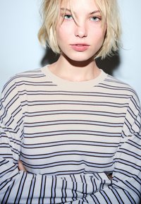 Młoda osoba z krótkimi blond włosami i niebieskimi oczami, ubrana w białą koszulę z długim rękawem w czarne i niebieskie poziome paski, z założonymi rękami.