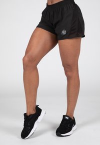 Zwarte sportshorts met een mesh zijpaneel, voorzien van een zilveren gorillalogo. Zwarte sportschoenen met witte zolen maken de look compleet.