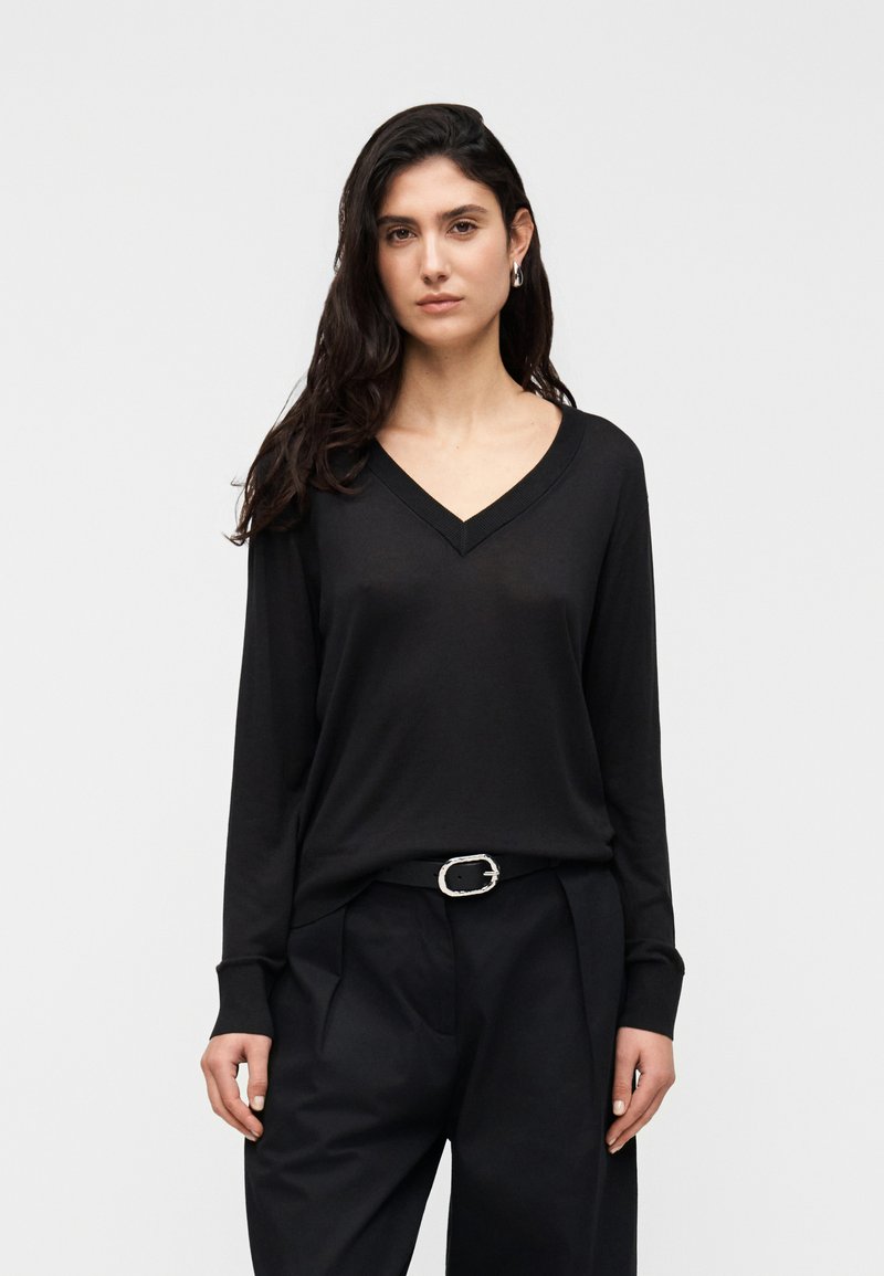 Haut noir à manches longues avec col en V, fabriqué en tissu doux, présentant une coupe décontractée et un détail c ôtelé le long de l'encolure. Associé à un pantalon taille haute.