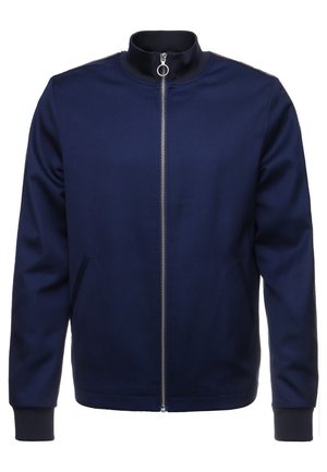 Marineblå lynlåstrøje med høj krave, sølvlynlås med cirkulært træk, lange ærmer og forlommer med slidser foran.