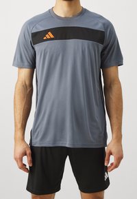 Šedá sportovní tričko s černým panelem a oranžovým logem, krátké rukávy, hladká textura, spárováno s černými šortkami s bílým logem.