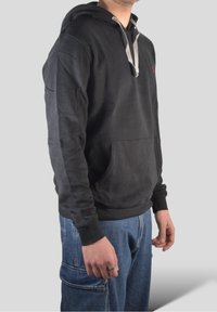 Czarna bluza z kapturem z przednią kieszenią, szarym sznurkiem i ściągaczami przy rękawach, noszona z niebieskimi dżinsami. Widok z boku.