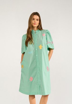 Žena nosi zeleno-biele pruhované košeľové šaty po kolená s krátkymi nadýchanými rukávmi a farebnými vyšívanými motívmi vtákov a kvetov.